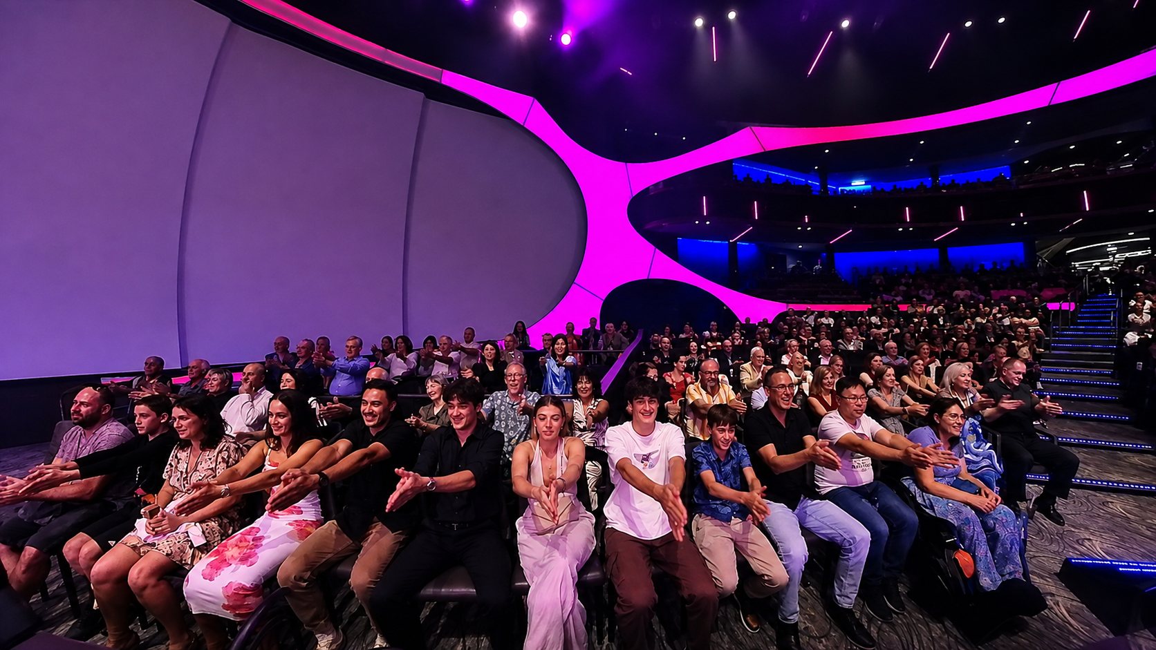 A full theatre lit in bright pink and blue, the whole audience with their hands locked together during the hypnosis routine
