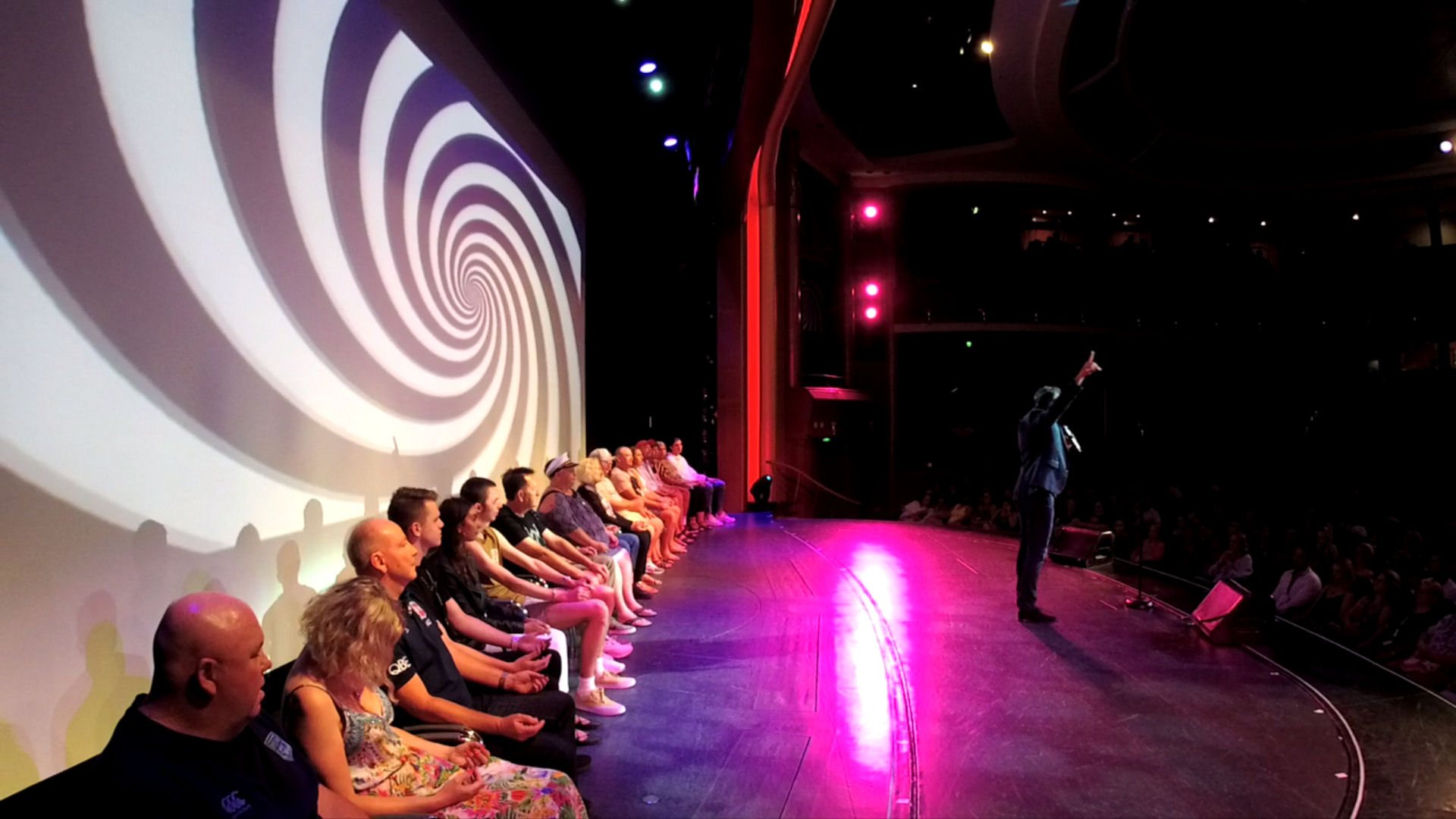 A row of hypnotised volunteers on a cruise-ship theatre stage, a huge spiral projected behind them